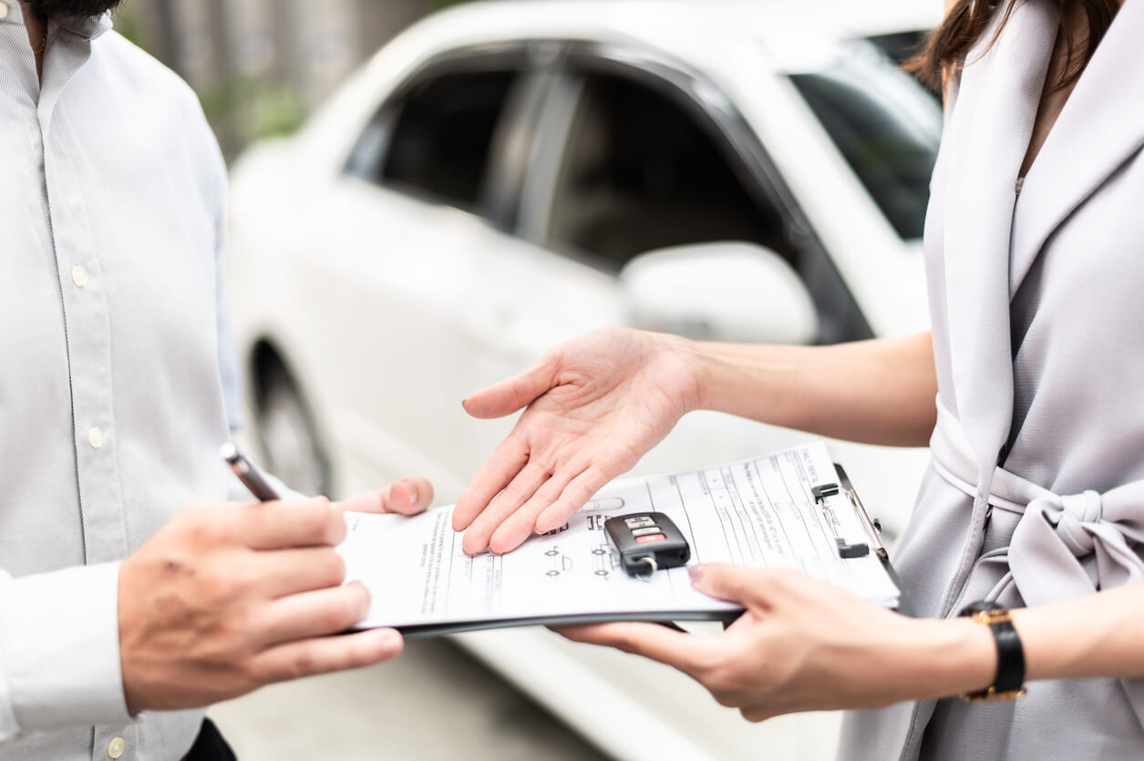 Two people reviewing vehicle paperwork with a car key resting on a clipboard in front of a white car